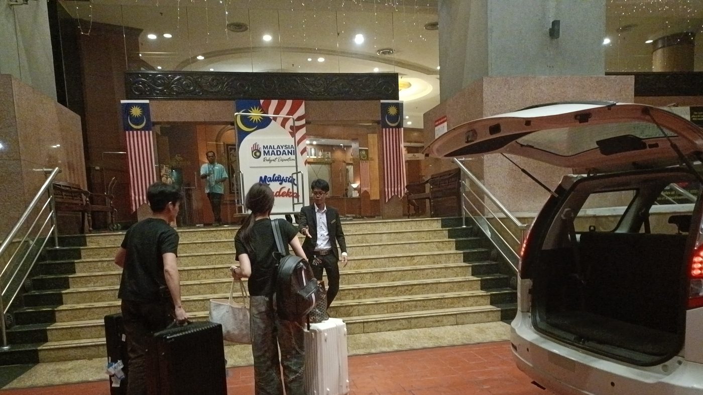Guests with roller bags arriving at a hotel lobby in Subang Jaya, with our white Proton Exora's boot open beside them